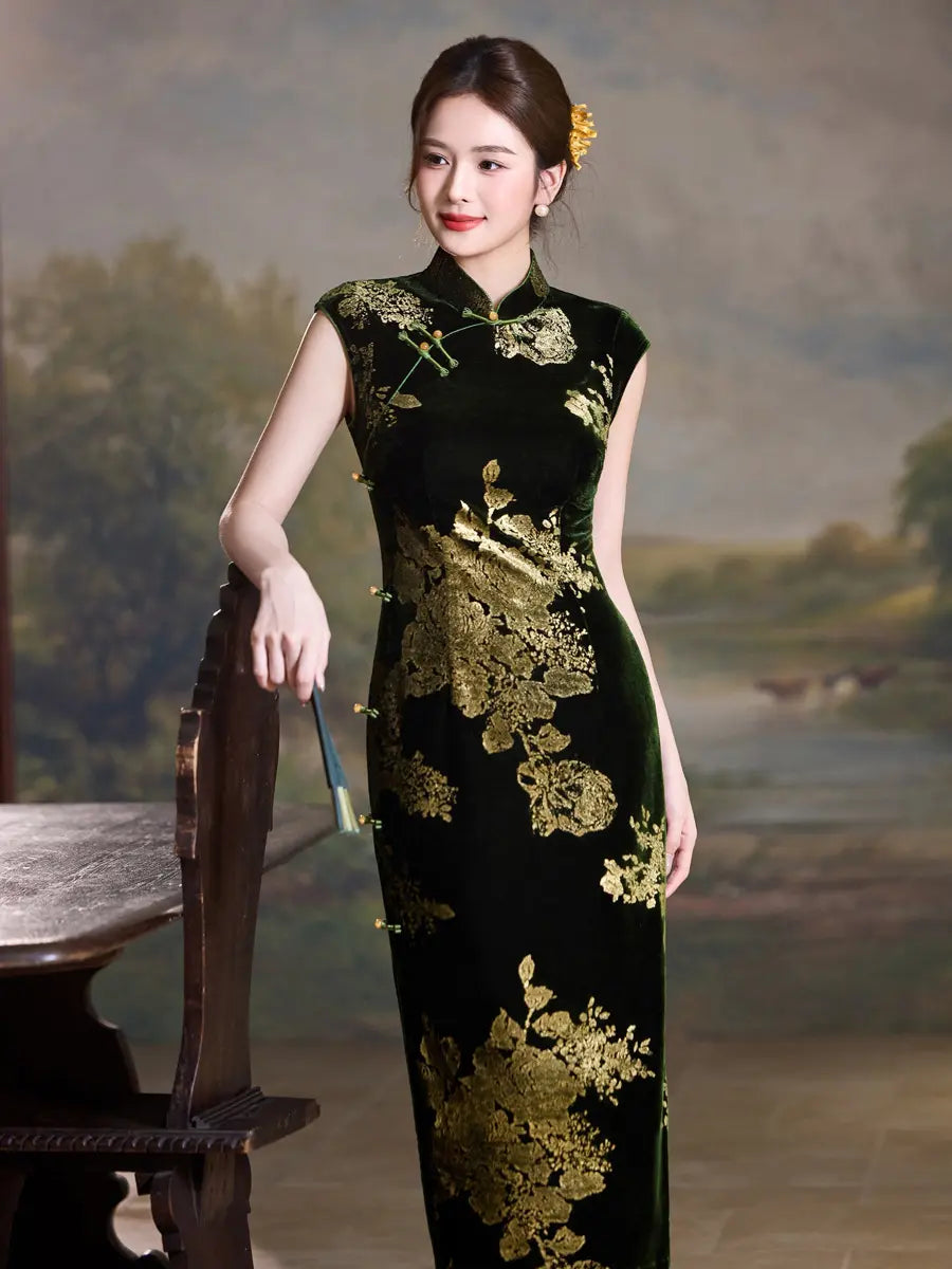 A woman models a sleeveless black velvet cheongsam-style dress with large gold floral embroidery, posed in front of a soft-focus painted landscape backdrop next to a wooden chair.