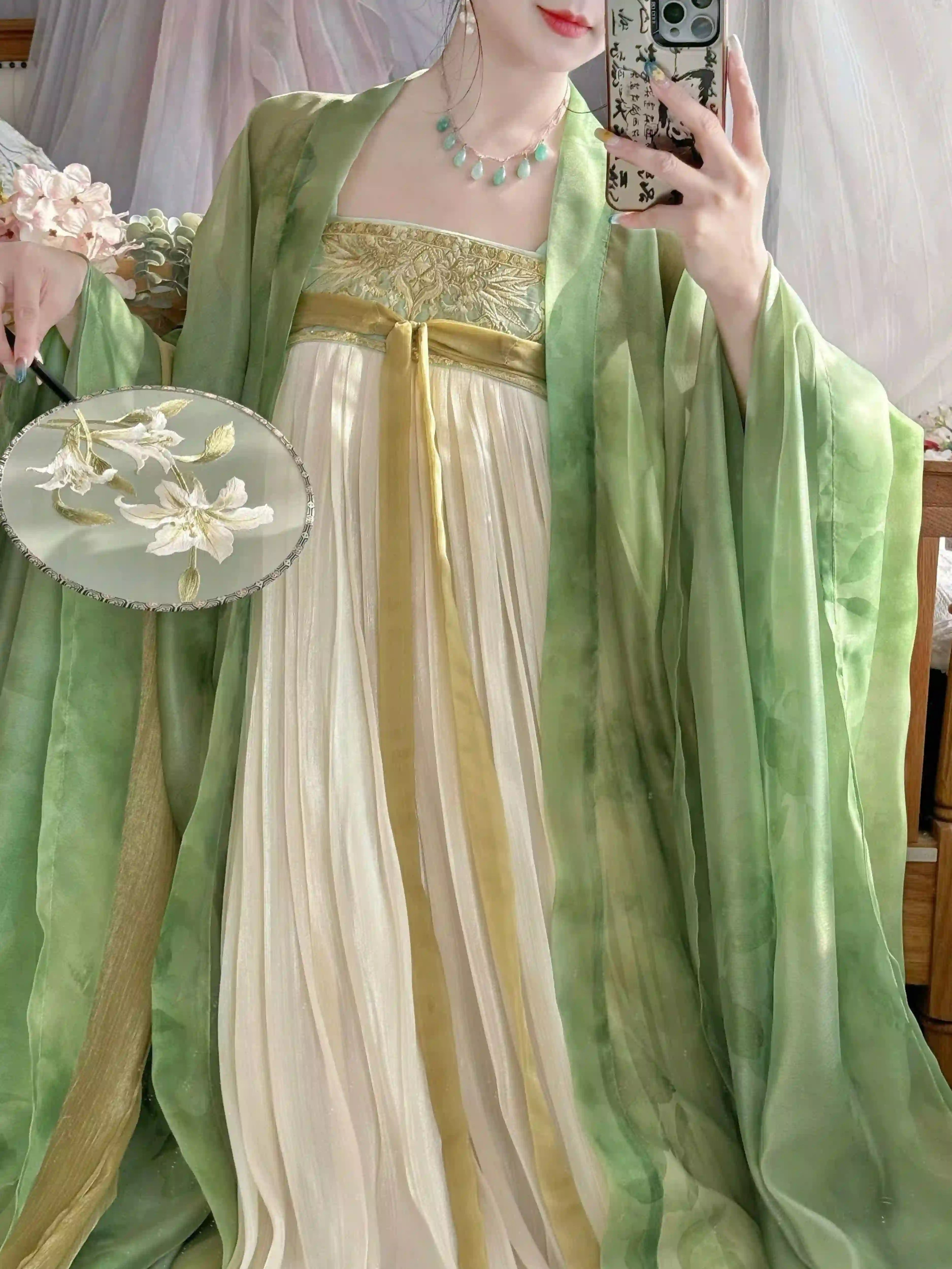 A person wearing a traditiona Chinesel-style tang dynasty green and cream hanfu dress with wide flowing sleeves, holding a floral round fan. The photo appears to be a mirror selfie in an indoor setting with soft pink drapery in the background.