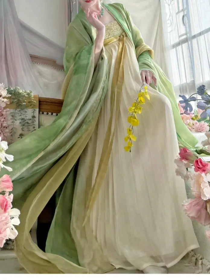 A woman wearing a flowing traditional tang dynasty Hanfu dress in soft sage green and cream, holding a small yellow flower sprig in a beautifully decorated indoor setting.