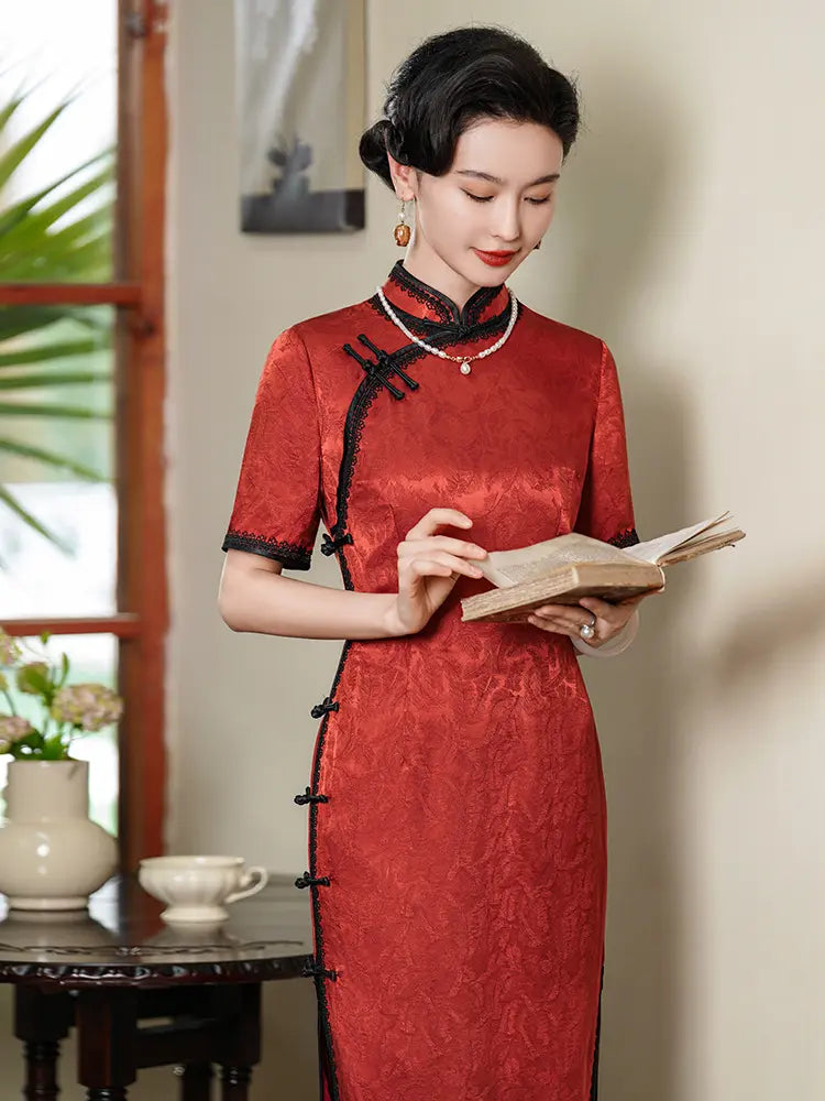Full-length shot of a woman in a deep crimson silk jacquard qipao with black lace trim and frog buttons, standing in a vintage-style room