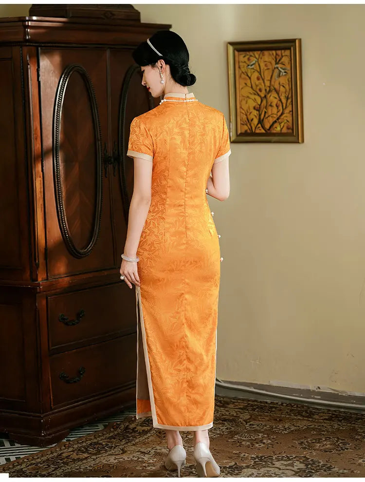 Woman in l Golden  orange Chinese  long qipao dress  standing in a room with a wooden cabinet and framed picture on the wall.
