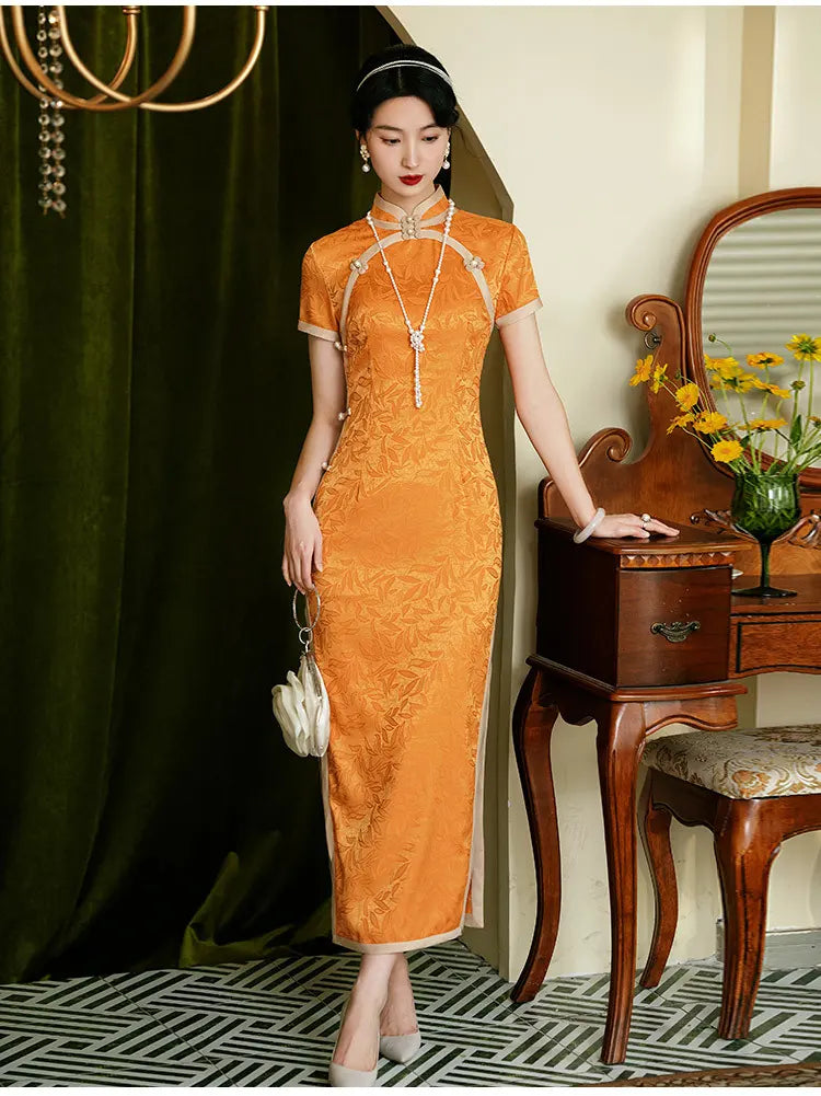 Woman in Golden  orange Chinese  long cheongsam dress standing in a room with wooden furniture and flowers.