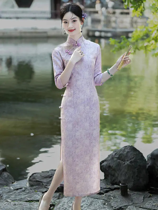 A woman wearing a light lavender-pink floral print cheongsam (qipao) dress stands by a scenic pond with rocks and green foliage in the background.