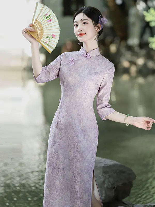 A woman wearing a lavender-purple traditional Chinese cheongsam dress with a subtle floral pattern, holding a decorative folding fan in a garden setting.