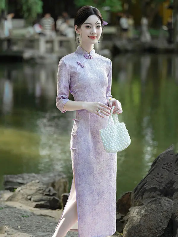 A woman wearing a light lavender-purple Chinese cheongsam (qipao) dress stands outdoors by a pond, holding a white woven handbag.