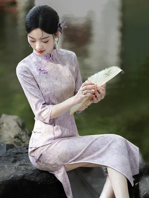 A woman wearing a light lavender Chinese cheongsam (qipao) dress sits on a stone in an outdoor garden setting, holding a folding fan.