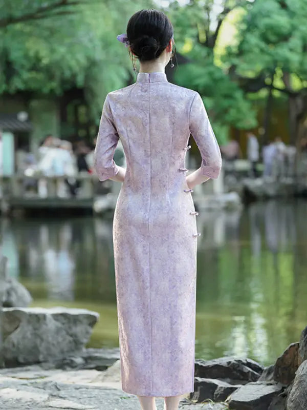 A woman stands with her back to the camera wearing a light pink floral-patterned cheongsam (qipao) dress in an outdoor garden setting with a pond and trees.