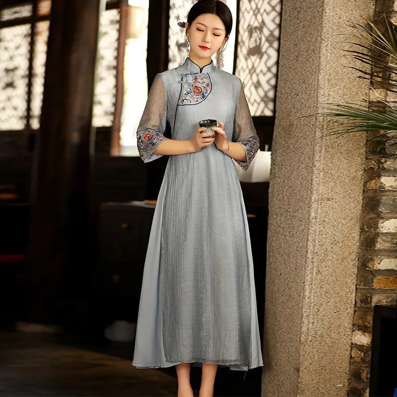 a chinese Woman in alight blue Modern A Line Loose Cheongsam Qipao Dress with Sheer Sleeve dress with floral embroidery holding a tea cup, standing against a stone wall.