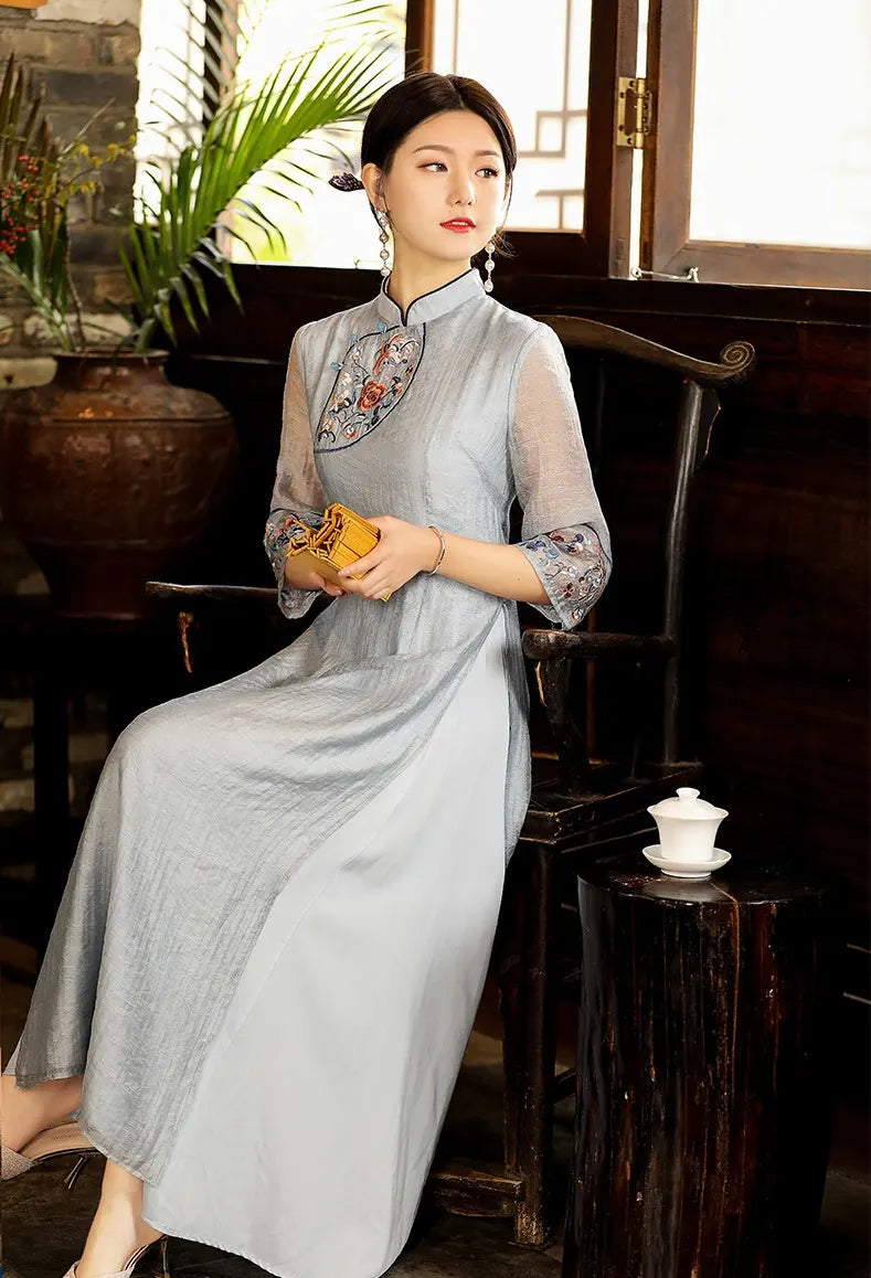 a chinese Woman in a light blue Modern A Line Loose Cheongsam Qipao Dress with Sheer Sleeve with floral embroidery sitting in a room with plants and a wooden stool.