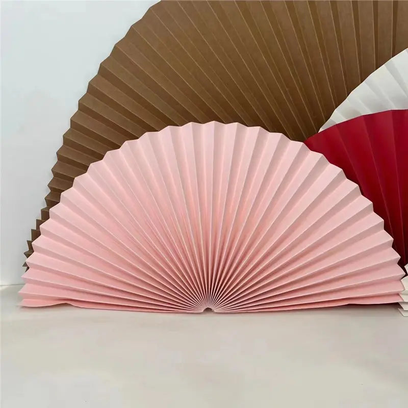Three pleated folding paper fans in pink, brown, and red, arranged in a fan-like stack against a plain light background.