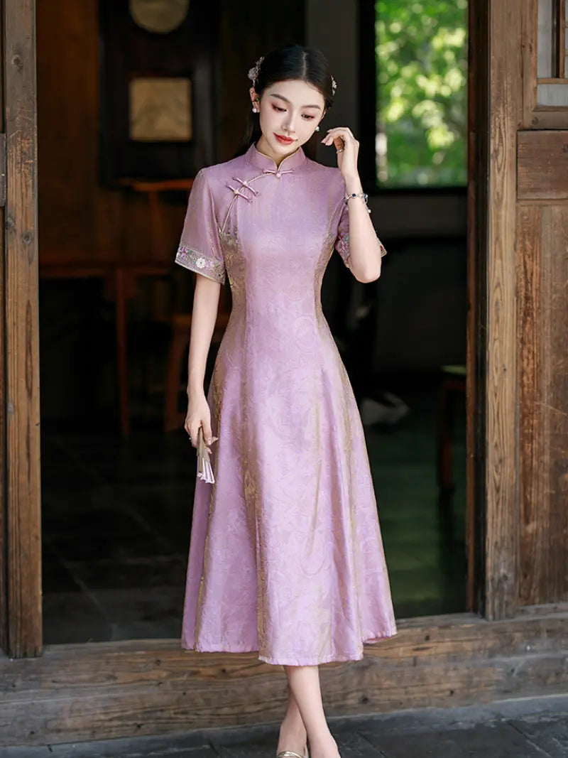 A woman wearing a lavender pink A-line qipao cheongsam dress with gold embroidery and lace trim, standing in a traditional wooden doorway.