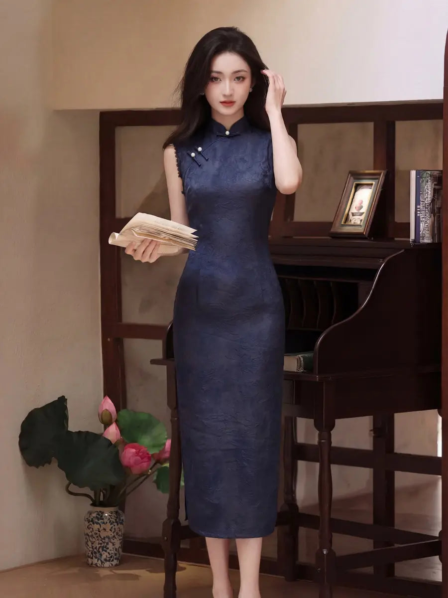A woman stands indoors wearing a sleeveless dark blue cheongsam-style dress with a high neck and side slit, holding a stack of papers. The setting includes a wooden cabinet, framed picture, and a vase with pink flowers.
