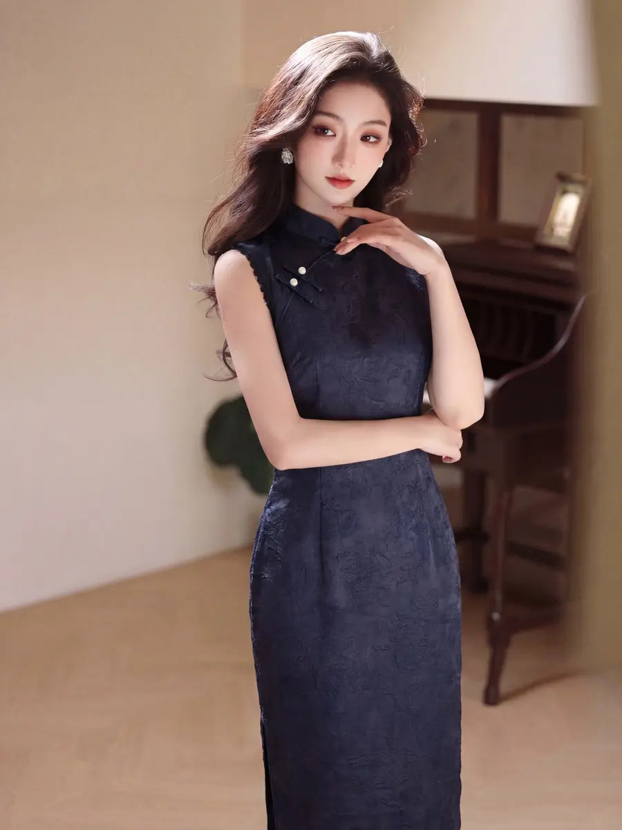 A woman wearing a sleeveless dark navy blue textured cheongsam dress, posing in a warmly lit indoor setting with a wooden piano in the background.
