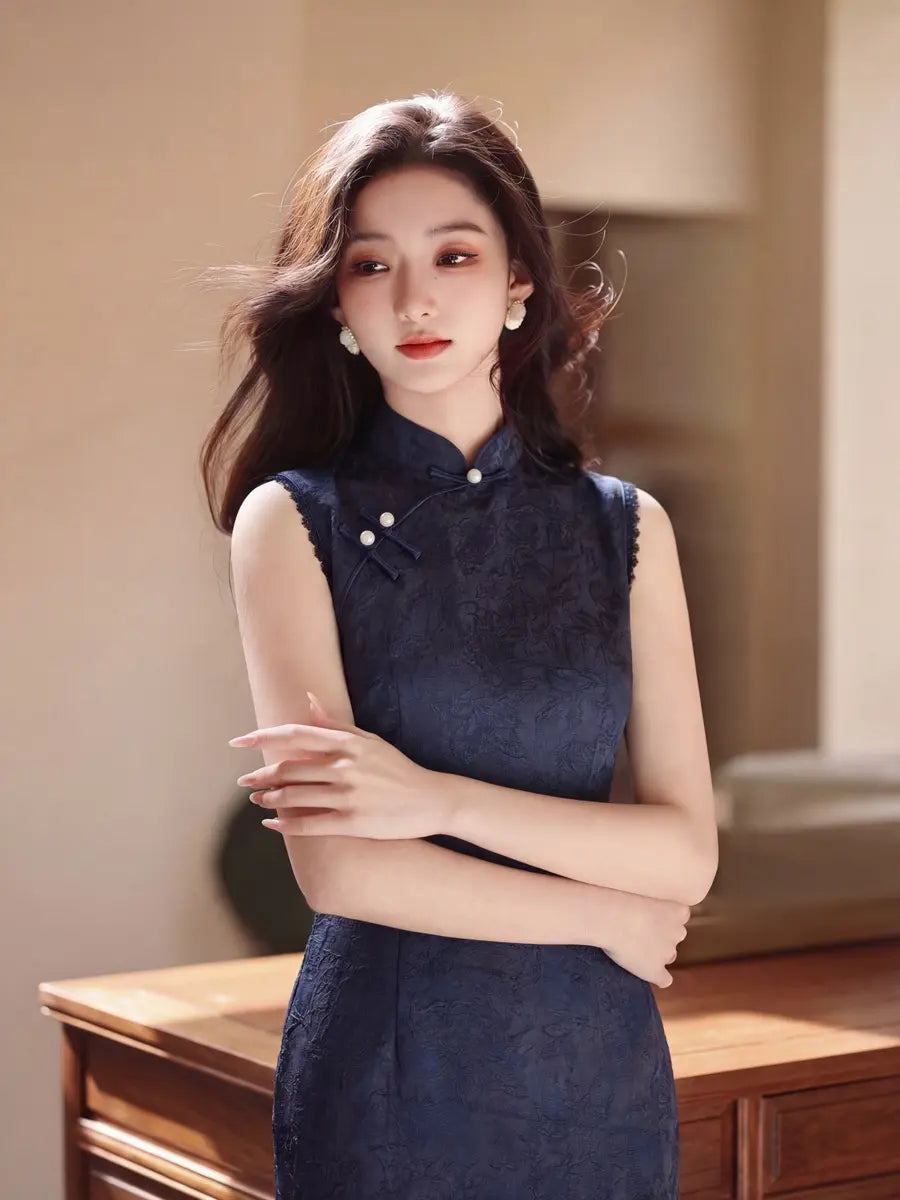A woman wearing a dark navy blue textured cheongsam dress, standing indoors with her arms crossed in front of a warm beige background and wooden furniture.