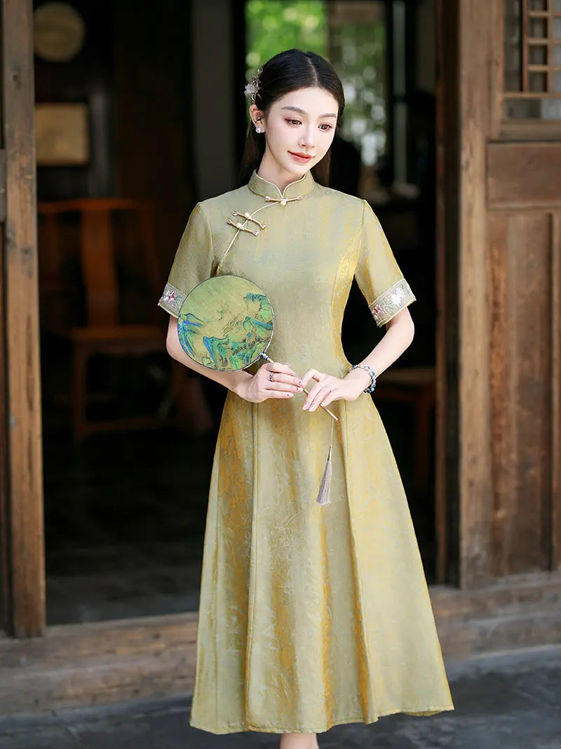A woman wearing a traditional Chinese cheongsam dress in a muted olive-gold color, holding a decorative round fan. She is standing in front of a rustic wooden doorway.
