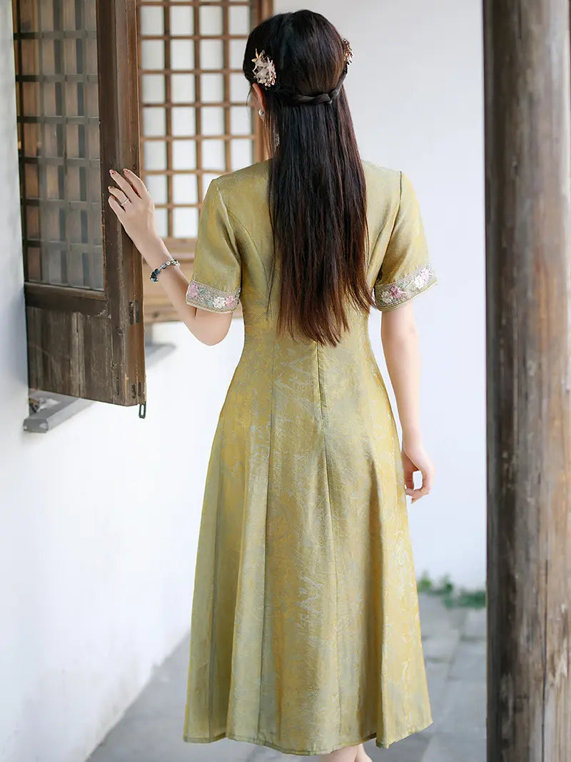 A woman viewed from behind wearing a short-sleeved, midi-length yellow-green A-line qipao cheongsam  dress with embroidered cuffs, standing in a traditional courtyard next to a wooden window.