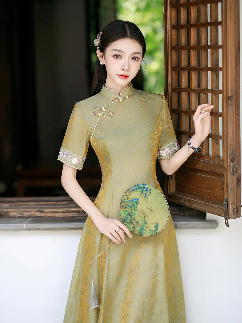 A woman wearing a traditional Chinese A-line cheongsam dress in olive green with gold embroidery and patterned sleeve cuffs, holding a round fan, standing in front of a wooden lattice window.