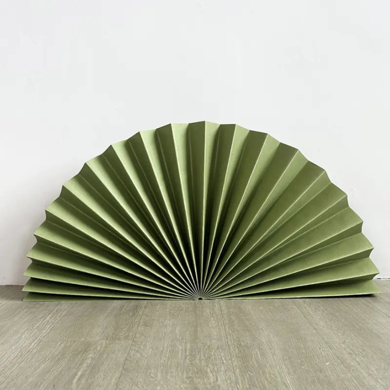 A large olive green pleated paper fan, partially unfolded, displayed on a light gray wood floor against a plain white wall.