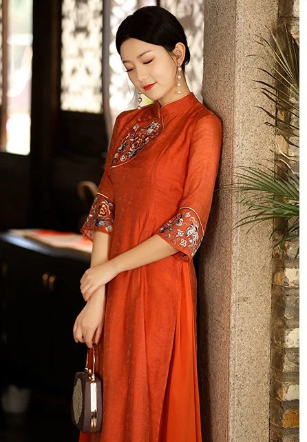 Woman in an orange traditional chinese  dress with floral embroidery leaning against a stone wall.