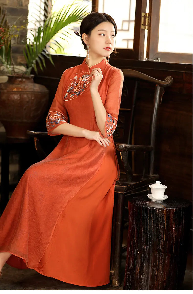 a chinese Woman in an orange Modern A Line Loose Cheongsam Qipao Dress with Sheer Sleeve sitting in a room with plants and a wooden chair.