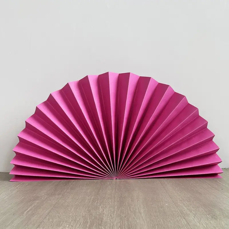 A bright pink pleated paper fan decoration, displayed open on a light gray wooden floor against a plain light gray wall.
