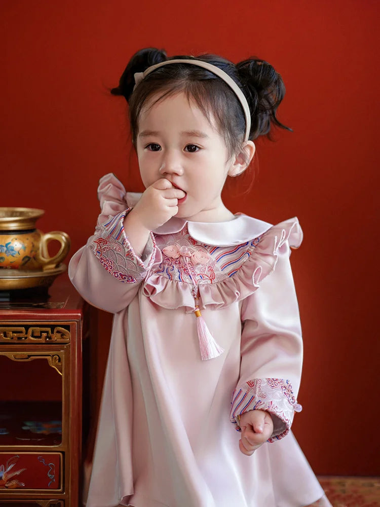 Young girl in a traditional pink Modern Chinese Baby Qipao Cheongsam Dress with Tassel outfit standing against a red wall.