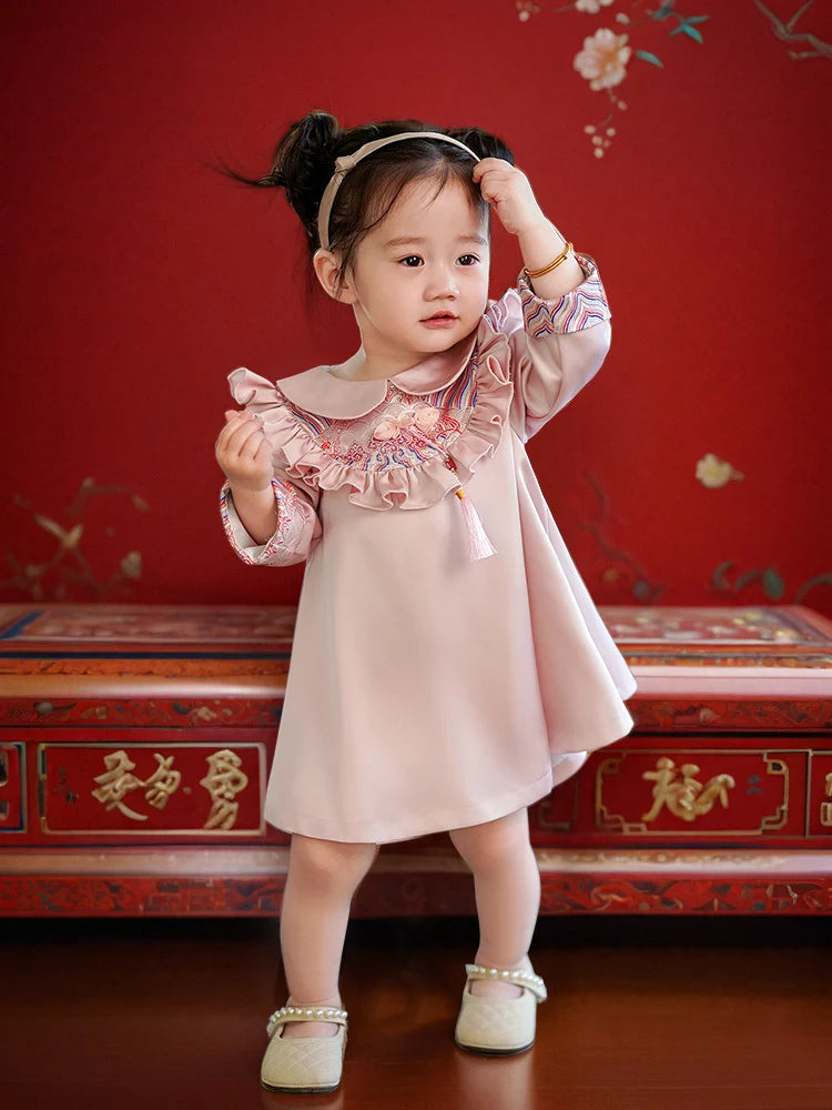 Young girl in a pink Modern Chinese Baby Qipao Cheongsam Dress with Tassel standing in front of a red background with floral patterns.