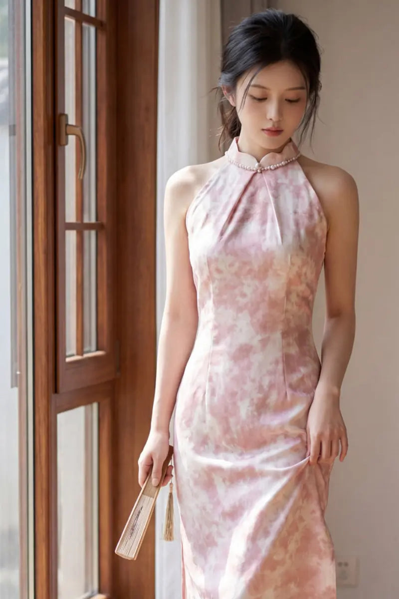 A woman wearing a sleeveless pink and beige watercolor-patterned  Modern Pearl-Embellished Halter-Style Qipao cheongsam dress, holding a small folding fan next to a wooden window frame.