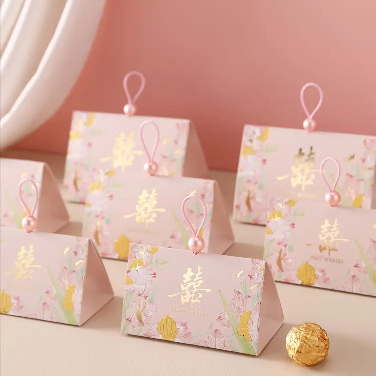 A set of pink Triangular Chinese  Double Happiness Wedding Favor Boxes with Pearl Loopwith floral prints and gold Chinese characters, each attached to a pink loop with a pearl bead, arranged on a beige surface against a soft pink background.