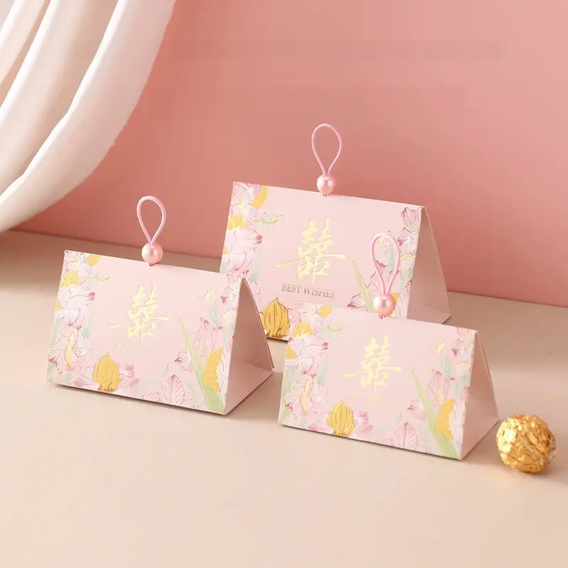Three pink tTriangular Chinese  Double Happiness Wedding Favor Boxes with Pearl Loop  with floral prints and gold Chinese characters, arranged on a beige surface against a soft pink background.