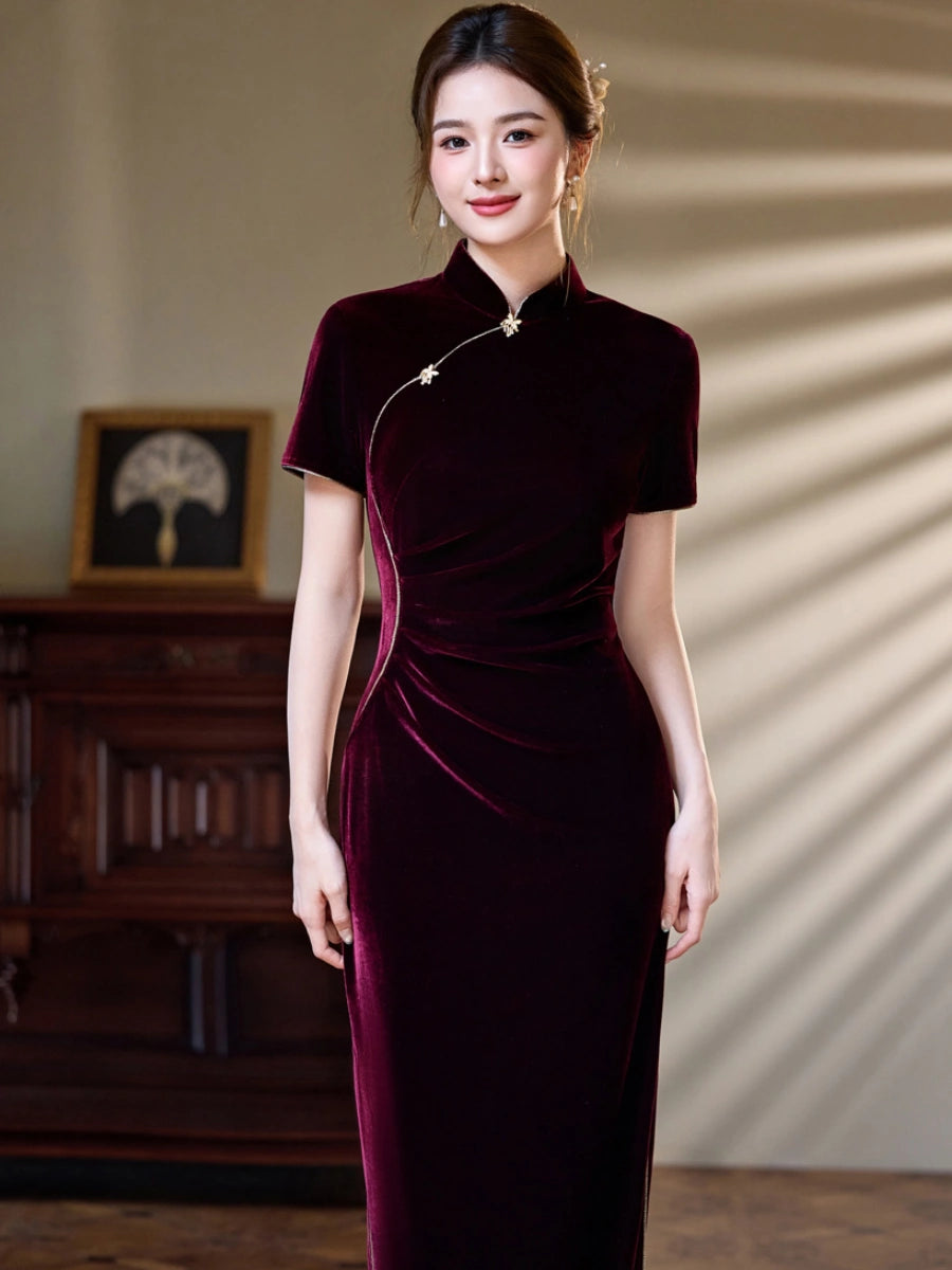 A woman wearing a floor-length, short-sleeved velvet cheongsam-style dress in deep burgundy, standing in an indoor setting with warm lighting and a wooden cabinet in the background.