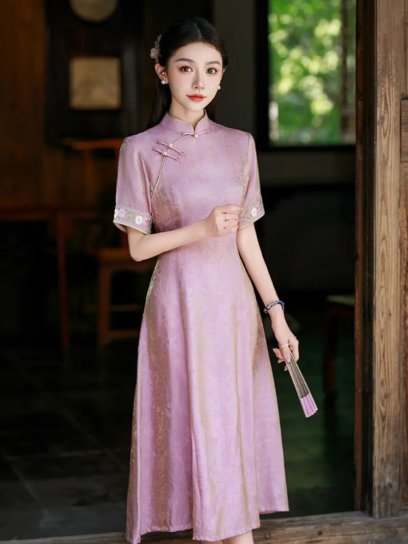 A woman wearing a light pink traditional  Chinese A-line qipao cheongsam dress with gold embroidery and silver-tone buttons, holding a matching pink folding fan in a rustic indoor setting.