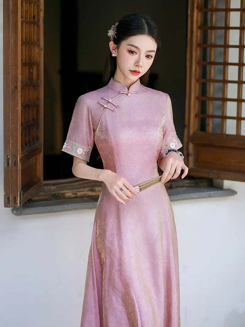 A woman wearing a light pink traditional Chinese cheongsam dress with embroidered sleeve cuffs, standing in front of a wooden window frame.
