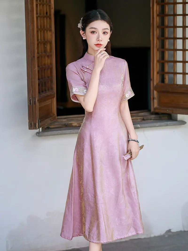 A woman wearing a light pink traditional Chinese cheongsam dress with gold embroidery and silver trim on the sleeve cuffs, standing in front of a white wall with wooden shutters.