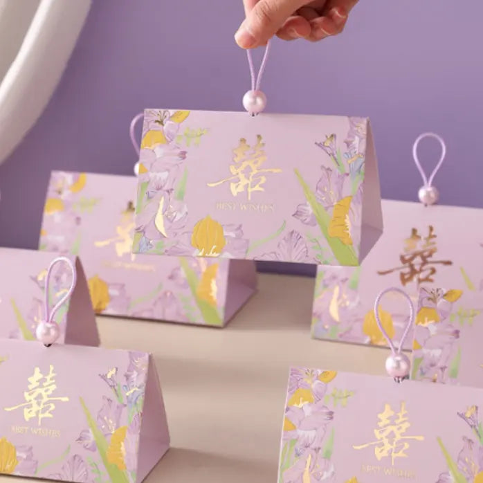A hand holds a lavender Triangular Chinese  Double Happiness Wedding Favor Boxes with Pearl Loop  with floral illustrations and gold Chinese characters. Several matching gift boxes are arranged on a tabletop against a soft lavender background.