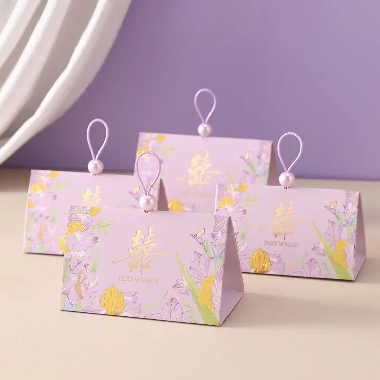 Four triangular Triangular Chinese  Double Happiness Wedding Favor Boxes with Pearl Loop s in pastel lavender with gold Chinese characters and floral illustrations, each with a pearl bead loop at the top, arranged on a beige surface against a purple and white background.