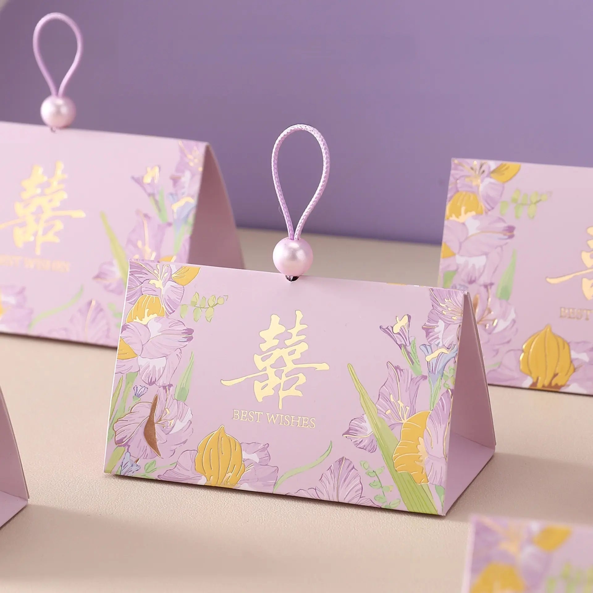 A lavender Triangular Chinese  Double Happiness Wedding Favor Boxes with Pearl Loop and pearl bead, decorated with floral illustrations and gold Chinese characters, displayed on a tabletop against a soft purple background.