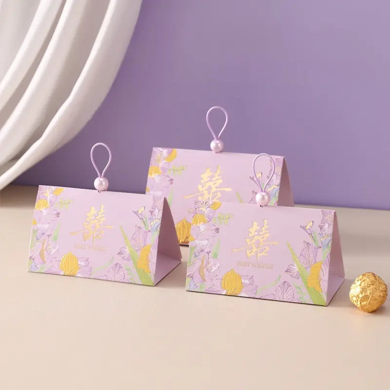 Three pastel lavender Triangular Chinese  Double Happiness Wedding Favor Boxes with Pearl Loop  with floral illustrations and gold Chinese characters, arranged on a beige tabletop against a lavender wall with a white draped curtain.