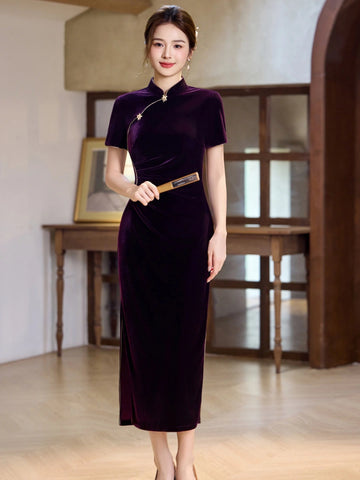 A woman wearing a dark plum velvet cheongsam midi dress with short sleeves, a high collar, and a silver-toned necklace, standing in a warm-toned indoor room with wooden furniture.