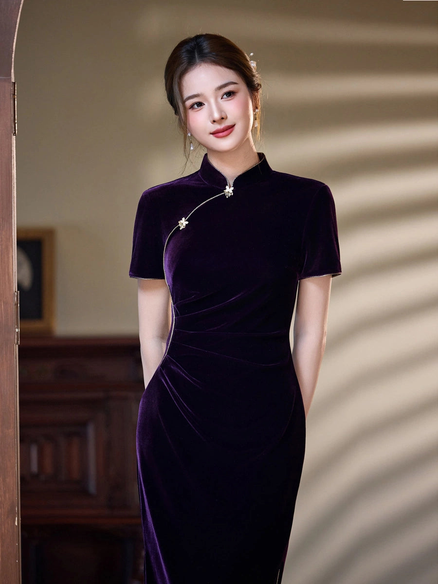 A woman wearing a short-sleeved, high-neck purple velvet cheongsam dress, standing in a warmly lit indoor setting with soft shadows on the wall.