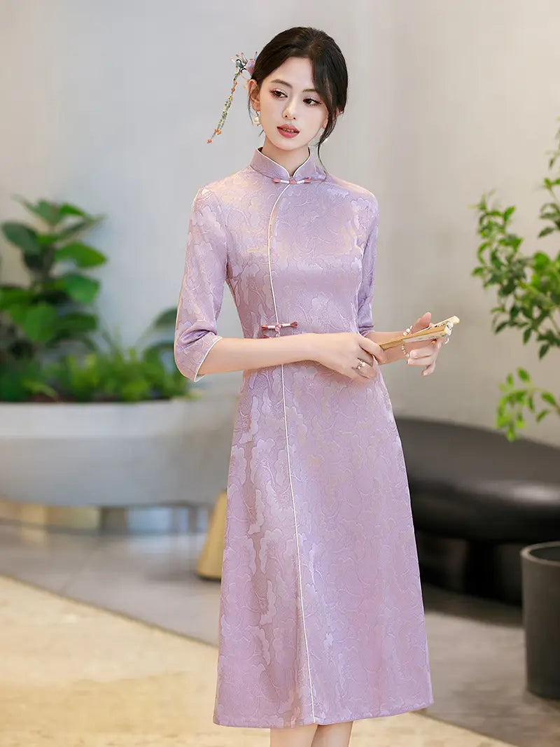 A woman wearing a lavender purple Chinese a line qipao cheongsam dress with three-quarter sleeves and a high collar, standing in a modern indoor setting with plants in the background.