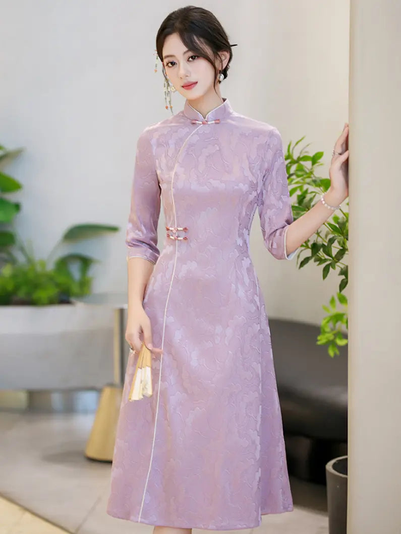 A woman wearing a lavender purple a line cheongsam dress with three-quarter sleeves and a high collar, standing in a modern indoor setting with plants in the background.