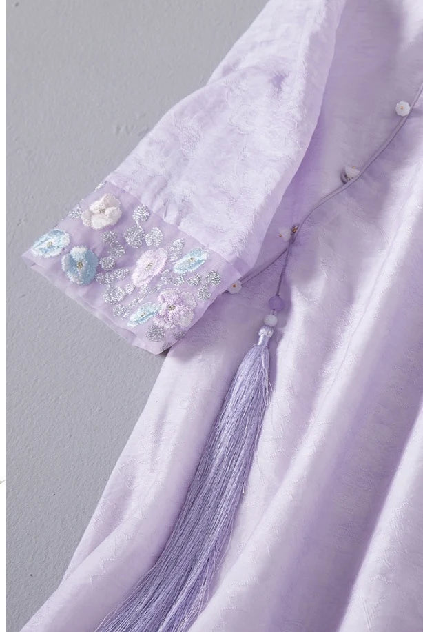 Close-up of a lavender garment with floral embroidery and tassel details on a gray background