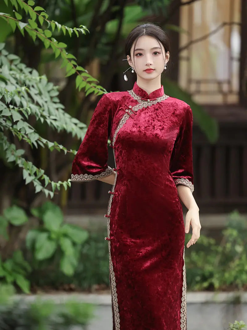 A woman wearing a deep red velvet cheongsam (qipao) dress with silver embroidered trim, standing in an outdoor garden setting with green foliage and a wooden structure in the background.