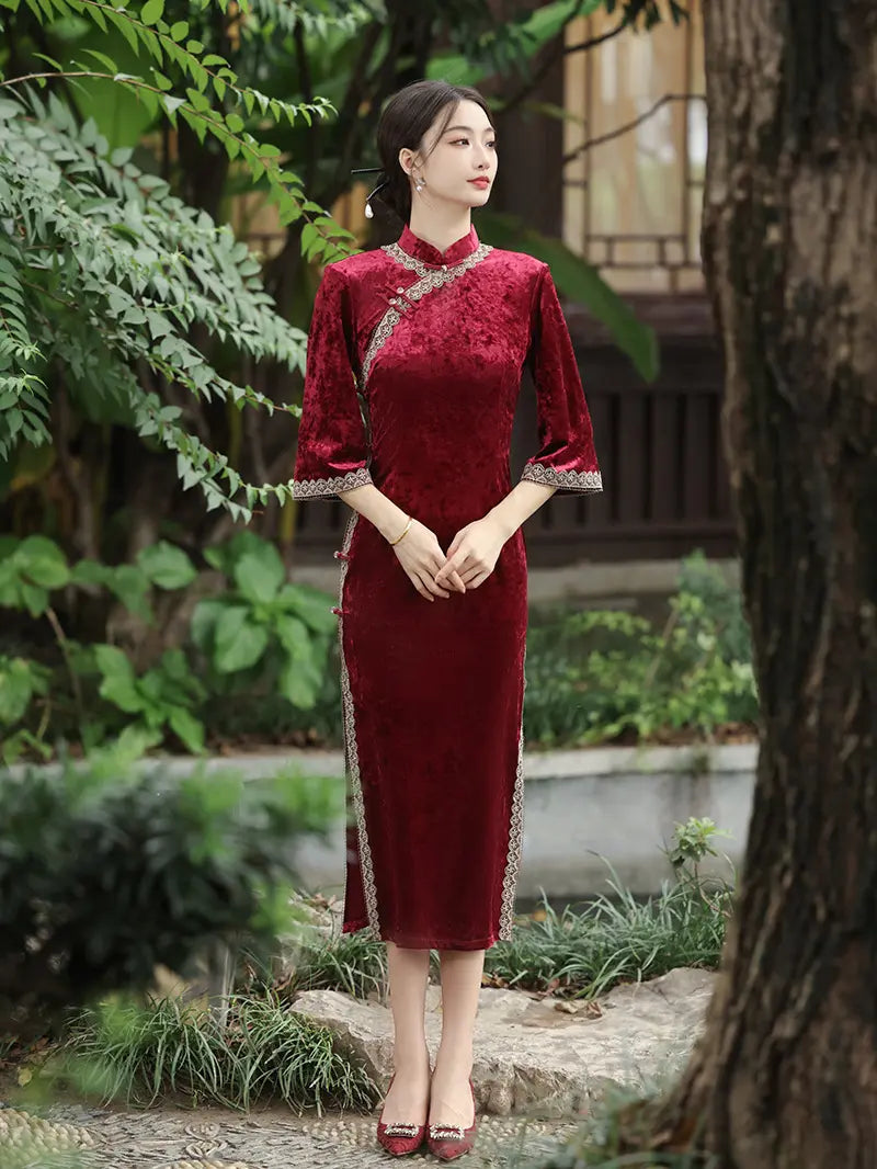 A woman wearing a deep red velvet cheongsam dress with silver embroidery and lace trim, standing in a garden setting with trees and foliage.