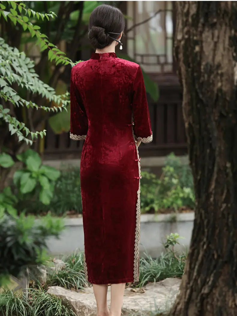 A woman viewed from behind wearing a deep red velvet cheongsam dress with gold lace trim at the cuffs and side, standing in a garden setting.