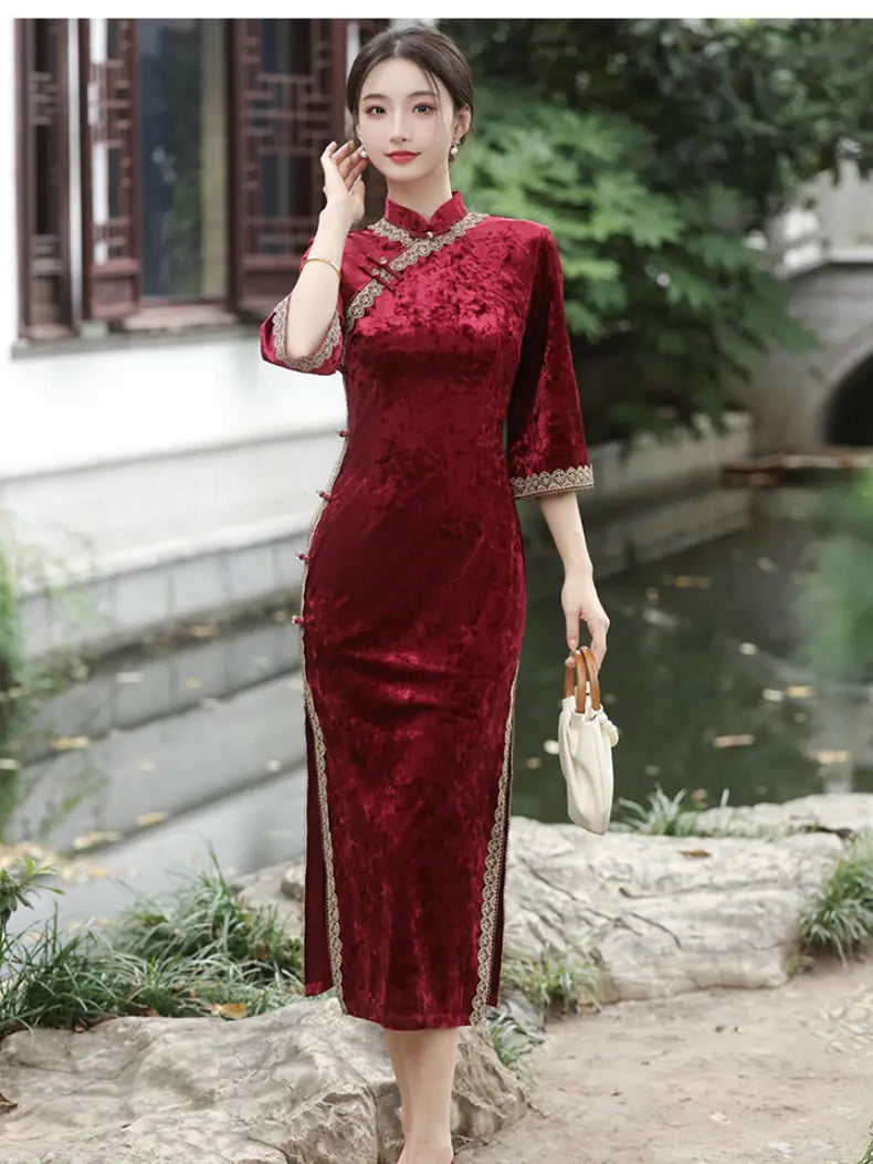 A woman wearing a deep red velvet cheongsam dress with gold embroidery and trim, standing outdoors near a stone path and water feature.