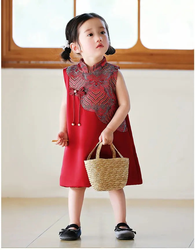 Child wearing a red and patterned Girls' "Good Fortune" Traditional Sleeveless A-Line Jacquard Qipao dress holding a straw bag indoors.