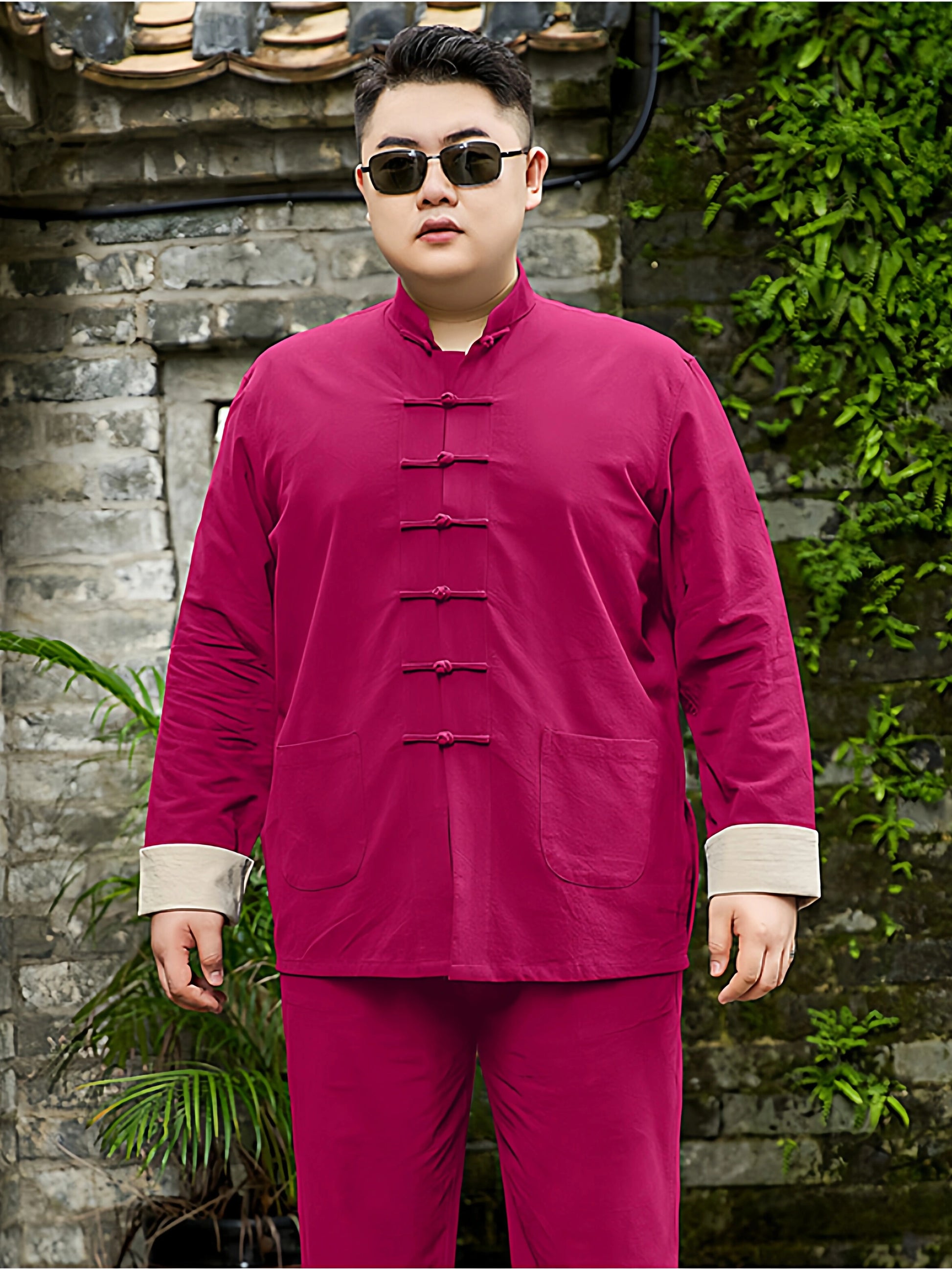 A man wearing a magenta-pink plus size traditional Chinese-style two-piece set with matching pants and sunglasses, standing in front of a stone wall with green plants.