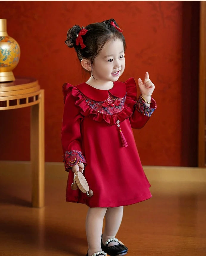 Young girl in a red Modern Chinese Baby Qipao Cheongsam Dress with Tassel  with ruffled collar and long sleeves, holding a stuffed toy, against an orange wall.